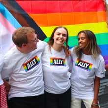 Load image into Gallery viewer, a group of three people wearing LGBTQIA+ tees with rainbow "ally" on them stand in front of a rainbow pride flag