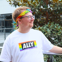 Load image into Gallery viewer, a person wearing a white tee shirt with LGBTQIA+ ally on it stands in front of a tree. they're wearing a rainbow bandana